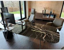 Modernes Büro mit einem Schreibtisch aus schwarzem Marmor mit weißen Aderungen. Im Hintergrund ein Holzregal mit Büchern und Dekorationen, daneben eine große Glasfensterfront mit Blick auf einen Garten.