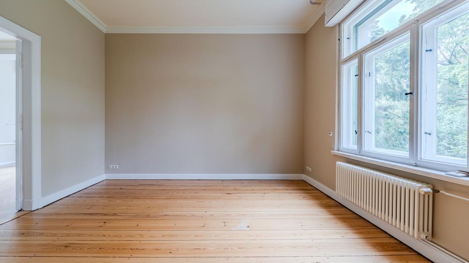 Unmöblierter Wohnraum mit hellem Holzdielenboden, beigefarbenen Wänden und weißer Decken- sowie Fußleisten. Rechts große, mehrteilige Fensterfront mit Blick ins Grüne und darunter ein weißer Heizkörper; links eine offene Türöffnung zu einem Nebenraum.
