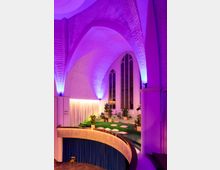 Innenansicht eines modern umgestalteten Kirchenraums mit hohen gotischen Fenstern, violettem Stimmungslicht und einem begrünten Podest mit Sitzkissen und Pflanzen. Im Vordergrund ist ein geschwungener Balkon mit blauen Vorhängen sichtbar.
