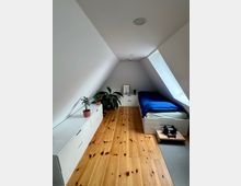 Schmales Dachzimmer mit schrägen weißen Wänden, Holzfußboden und einem Einzelbett mit blauem Bettbezug neben einem großen Dachfenster. Entlang der linken Seite stehen weiße Kommoden mit Topfpflanzen, am Ende des Raums eine weitere niedrige Kommode.