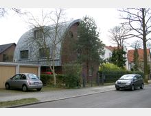 Straßenansicht in einem Wohngebiet mit einem modernen Wohnhaus, das durch seine gebogene Metallfassade und rotbraune Ziegelwände auffällt. Im Vordergrund stehen zwei parkende Autos vor einem Carport und einige Bäume säumen das Grundstück.