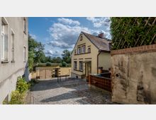 Gepflasterte Gasse mit einem gelb gestrichenen Haus mit dunkelbraunen Fensterrahmen und einem kleinen Vordach, umgeben von einer Mauer und Bepflanzungen; im Hintergrund befinden sich Bäume und ein blauer Himmel mit Wolken.