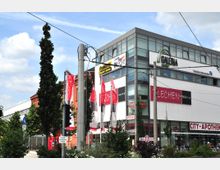 Moderne Geschäftsgebäude mit Glasfassade und verschiedenen Geschäften, darunter eine Apotheke, neben einem roten Backsteingebäude; im Vordergrund Bäume, Fahrräder und Straßenbahnschienen.