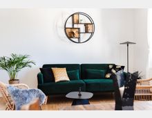 Wohnzimmer mit einer dunkelgrünen Samt-Couch, dekoriert mit Kissen in Gold- und Schwarztönen. An der weißen Wand darüber hängt ein rundes, modernes Regal mit Deko-Objekten, links steht eine grüne Zimmerpflanze und rechts eine schlichte Stehlampe.