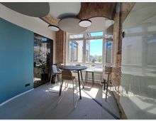 Moderner Besprechungsraum mit einer Mischung aus Backstein- und glatten Wänden, blauer Akzentwand und einer Glastrennwand. Ein rundes Hochtisch-Set mit Stühlen steht vor großen Fenstern, die einen Blick auf umliegende Gebäude und blauen Himmel bieten.