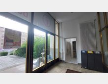 Eingangsbereich eines Gebäudes mit großen Glastüren, durch die eine Terrasse mit grüner Vegetation sichtbar ist. Rechts befinden sich Abfalltrennbehälter, ein Heizkörper und eine offene Tür, die in einen Flur mit gefliesten Wänden führt.