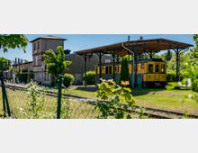 Kleiner, ländlicher Bahnhof mit Gleisen im Vordergrund, einem älteren Stationsgebäude und Signaltechnik. Rechts steht unter einem überdachten Bahnsteig mit gusseisernen Stützen ein gelber historischer Triebwagen, umgeben von Grünflächen und Bäumen.