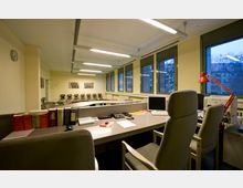 Ein Büroraum mit modernen Arbeitsplätzen, mehreren Schreibtischen, Bürostühlen und technischen Geräten wie Computern und Telefonen. Im Hintergrund befinden sich große Fenster, eine Reihe von Stühlen entlang der Wand sowie minimalistische Dekoration mit Wandbildern.