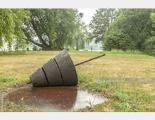 Skulptur eines stilisierten, horizontal liegenden Bohrkopfes aus mehreren Segmenten und Metallelement, auf einer Wiese in einem parkähnlichen Gelände mit Bäumen im Hintergrund, nahe einem Gewässer.