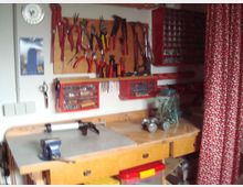 Werkstatt mit einer hölzernen Werkbank, darüber eine Wand mit Werkzeugen wie Zangen und Schraubendrehern, die auf einem Holzbrett aufgehängt sind. Daneben befinden sich rote Schraubenkästen und ein Regal mit Schraubenfächern; ein Teil des Raums ist durch einen gemusterten Vorhang abgetrennt.