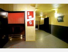 Flur eines Kinos mit gelb-beigen Wänden und schwarzen Details, der zu zwei Sälen führt, markiert mit den Buchstaben „A“ und „B“. Der Saal „B“ ist links erkennbar, mit geöffnetem Türrahmen, roten Kinositzen und einer roten Wand im Inneren.