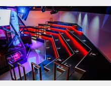 Futuristischer Innenraum mit einer Bühnen- oder Sitzanordnung aus schwarzen Podesten, die mit roten LED-Streifen beleuchtet sind. Im Vordergrund befinden sich metallene Geländer, und an der Seite sind weiße Wände und bunte Projektionslichter erkennbar.