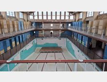Ein leeres, verfallenes Schwimmbad mit türkisfarbenen Fliesen und verblassten Wänden. Auf zwei Ebenen befinden sich Umkleidekabinen mit blauen und gelben Türen sowie eine Galerie mit Metallgeländern. Große Fenster lassen Tageslicht in den Raum.