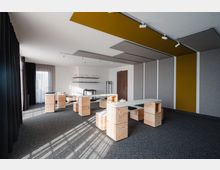 Heller, moderner Seminarraum mit modularen Holztischen und gepolsterten Sitzelementen. Der Raum verfügt über grauen Teppichboden, Akustikpaneele an Wänden und Decke sowie große Fenster mit dunklen Vorhängen, die Tageslicht einlassen.