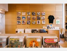 Innenraum eines Plattenladens mit Holzverkleidung, Regalen voller Schallplatten und einer Wand aus Lochplatten, an der Schallplattencover, ein schwarzes T-Shirt und eine weiße Tasche hängen. Im Vordergrund sind Schallplatten in Fächern arrangiert, während im Hintergrund ein runder Tisch mit einer blauen Lampe und einem Laptop steht.