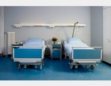 Zwei Krankenhausbetten mit blauen Polstern stehen nebeneinander in einem hell beleuchteten Patientenzimmer mit blauem Boden und grauen Wänden. Zwischen den Betten befindet sich ein Nachttisch mit Blumen, darüber ist eine Leiste mit Steckdosen und Leselampen angebracht.