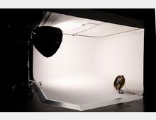 Ein Fotostudio mit einem weißen Hintergrund, einem großen schwarzen Studiolicht auf einem Stativ links und einem gelben Ventilator am Boden rechts. Der Raum ist spärlich beleuchtet, mit einer hohen, sichtbaren Decke und dunklen Flächen im Vordergrund.
