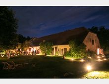 Ein großer, beleuchteter Hof bei Nacht mit einem länglichen Gebäude aus Stein und Ziegeldach; im Vordergrund eine Wiese mit Laternen und Gartenmöbeln, umgeben von Bäumen und Sträuchern. Im Hintergrund sind Menschen bei einem Fest oder einer Veranstaltung nahe dem Gebäude zu erkennen.