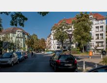 Straßenszene in einem Wohngebiet mit mehrstöckigen Altbauten im Stil des frühen 20. Jahrhunderts, umgeben von Bäumen. Geparkte Autos säumen die Straße, während ein Verkehrsschild und eine orangefarbene Mülltonne den Gehweg begrenzen.