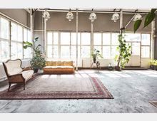 Industriehalle mit großen Fenstern, die viel Tageslicht einlassen. Im Vordergrund befinden sich ein bunter Teppich, zwei Vintage-Sofas, mehrere Pflanzen und eine Tür auf der rechten Seite.