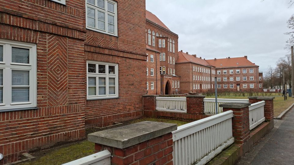 Backstein-Schulgebäude mit weiß gerahmten Fenstern und einem spitzbogigen Eingangsportal, umgeben von niedrigen Ziegelmauern mit weißen Geländern. Im Hintergrund stehen weitere langgestreckte Backsteinbauten mit roten Ziegeldächern entlang eines Gehwegs und einer Grünfläche.