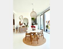 Heller Essbereich in einem modernen Interieur mit einem runden Holztisch, vier Holzstühlen und einem runden Juteteppich. Im Hintergrund steht eine Holzkommode mit einer Pflanzendekoration, und große Glasfenster bieten Blick auf Gebäude draußen.