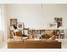Wohnzimmer mit minimalistischer Einrichtung, bestehend aus beigen Polstersesseln und einem Sofa. Im Hintergrund stehen niedrige Regale mit Büchern, darauf befinden sich gerahmte Kunstwerke und Poster sowie ein Plattenspieler; eine einzelne Glühbirne hängt von der Decke.