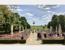 Eine Terrasse mit Biergarten, ausgestattet mit rot-weiß karierten Tischen und Bierbänken, umgeben von großen grünen Bäumen und Sonnenschirmen. Im Hintergrund ist eine weite, offene Landschaft mit Feldern und blauem Himmel mit vereinzelten Wolken sichtbar.