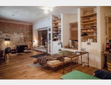 Ein gemütlich gestalteter Wohnraum mit einer Mischung aus modernem und rustikalem Stil. Sichtbare Putzwände, Holzboden, Regale mit Gegenständen, eine Schlafliege mit Decke und Kopfkissen sowie verschiedene Sitzmöbel prägen den Raum; eine Glastür mit Vorhang und diverse Lampen schaffen eine warme Atmosphäre.