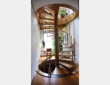 Holz-Spiraltreppe in einem hellen Innenraum mit weißen Wänden, umgeben von Pflanzen und wandhängenden Bildern. Im Hintergrund steht ein Tisch mit Stuhl vor einem Fenster, das Tageslicht hereinlässt.