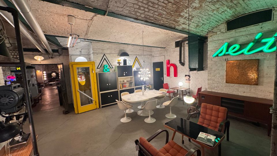 Ein Raum mit Gewölbedecke und freiliegenden weißen Ziegelwänden, eingerichtet im Retro-Stil. Zu sehen sind eine gelbe Telefonzelle, Neon-Schriftzüge, Designermöbel mit geschwungenen Formen sowie Regale und Lampen im Vintage-Look.