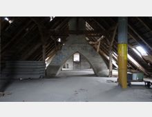 Dachboden eines alten Gebäudes mit sichtbaren Holzsparren, einem gemauerten, bogenförmigen Stützpfeiler in der Mitte und teils verblasster Graffiti auf den Wänden. Der Raum ist spärlich eingerichtet, mit Bauschutt, Metallplatten und einem gelben Rohr an einer Seite.