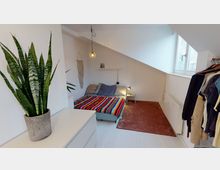 Schlafzimmer mit schräger Decke, einem Bett mit bunter Tagesdecke, einer Nachttischlampe und einem kleinen Beistelltisch. Im Vordergrund eine Pflanze auf einer weißen Kommode, rechts ein offener Kleiderständer; Tageslicht kommt durch ein Fenster mit weißem Rahmen.