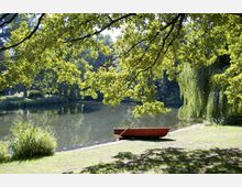 Ein ruhiger Park mit einem See im Hintergrund, umgeben von Bäumen und dichtem Grün. Am Ufer liegt ein rotes Ruderboot, teilweise im Schatten von überhängenden Ästen einer Weide.
