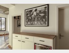 Helle, moderne Innenraumaufnahme eines Wohn- oder Arbeitsbereichs mit einem weißen Sideboard und Holzarbeitsfläche im Vordergrund. An der Wand hängt ein großes gerahmtes Schwarz-Weiß-Bild, im Hintergrund sind ein großes Fenster, ein Tisch und ein Türdurchgang sichtbar.