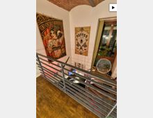 Innenansicht eines Tattoo-Studios mit hoher, weißer Wand und gewölbter Ziegeldecke, gesehen von einer Galerie mit Metallgeländer und Holzboden. An der Wand hängen große Tattoo-Poster, daneben ein hohes Fenster; darunter ist ein Arbeitsplatzbereich mit Schreibtisch und Ausstattung erkennbar.