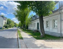 Straßenansicht einer ruhigen Wohngegend mit einer schmalen Straße, flankiert von Gehwegen und großen grünen Bäumen. Auf der rechten Seite befindet sich ein weißes, einstöckiges Gebäude mit modernen Fenstern und einer dunkelgrünen Eingangstür.