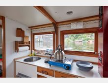 Innenraum einer kleinen Küche auf einem Hausboot mit Blick auf das Wasser durch zwei große Fenster mit Holzrahmen. Zu sehen sind eine Spüle, ein blauer Campingkocher mit Metallkessel, eine Kaffeemaschine aus Glas und gestapelte Teller, Umgeben von weißem Mobiliar und Holzverkleidung. Im Hintergrund sind Uferbebauung und Vegetation sichtbar.