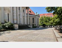 Historisches Gebäude mit massiven Säulen und roten Dachziegeln, umgeben von einem gepflasterten Platz mit Bäumen und Sitzbänken. Im Hintergrund sind weitere Gebäude und spazierende Personen erkennbar.