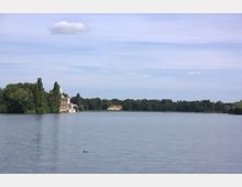 Seeufer mit einem roten Backsteingebäude auf der linken Seite, umgeben von Bäumen, und in der Ferne ein weiteres Gebäude mit rotem Dach. Der See dominiert das Bild, im Vordergrund schwimmt ein Vogel auf dem Wasser.