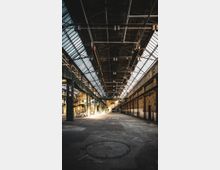 Weites Inneres einer Industriehalle mit hohen Ziegelwänden, einer verglasten Dachkonstruktion und sichtbarem Stahlträgerwerk. Der Boden besteht aus Beton, und seitlich sind metallene Treppen sowie Fenster und Türen zu sehen.