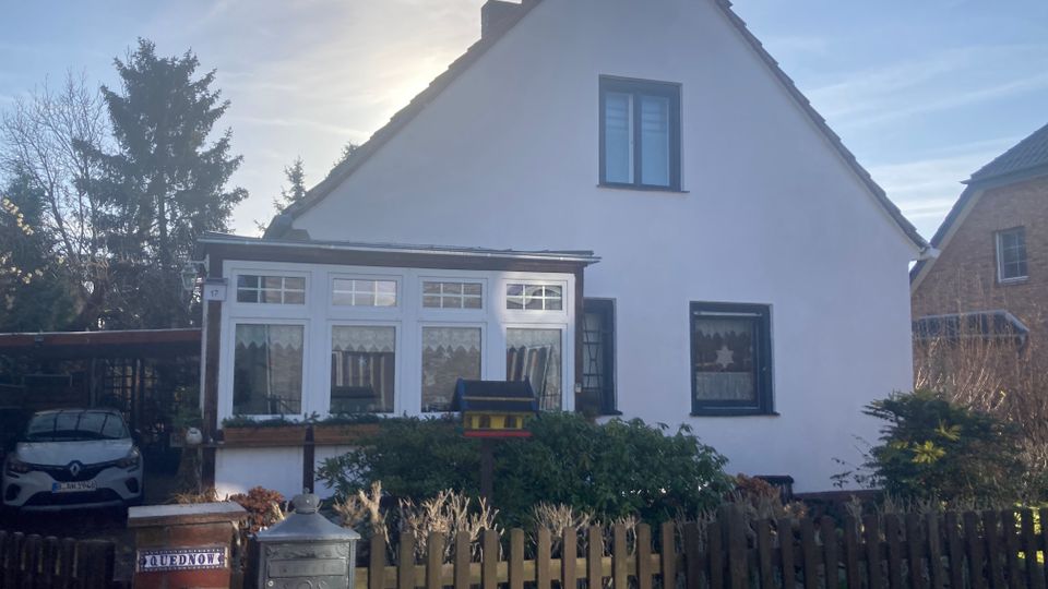 Ein weißes Einfamilienhaus mit Satteldach, einem Wintergarten mit großen Fenstern im Vordergrund und einem hölzernen Zaun, der das Grundstück umgibt. Im Garten befinden sich grüner Strauchbewuchs, und links neben dem Haus steht ein überdachter Carport mit einem geparkten Auto.