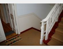 Treppenhaus mit weißem Holzgeländer und roten Stufen, die mit einem beigen Teppichläufer versehen sind. Im Hintergrund ist eine geöffnete Tür zu einem angrenzenden Raum mit Holzboden sichtbar.