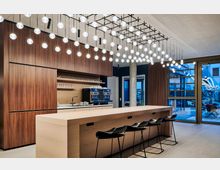 Moderne Küche mit hölzernen Schränken und einer großen Holztheke, umgeben von schwarzen Barhockern. Vielzahl an hängenden Kugelleuchten an der Decke, daneben bodentiefe Fenster mit Blick auf einen Außenbereich mit Sitzgelegenheiten.