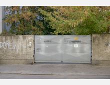 Graues Metalltor mit der Aufschrift „Parkhof“ und „Augustinenhof“, flankiert von einer grauen, mit Graffiti besprühten Betonmauer. Im Hintergrund sind Bäume mit teils herbstgefärbtem Laub zu sehen.