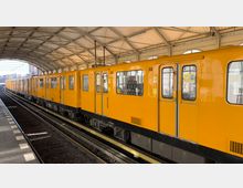 Gelber U-Bahn-Zug an einem überdachten Bahnsteig mit gewölbtem Dach und Oberlichtern, Gleise und Bahnsteigkanten deutlich sichtbar. Im Hintergrund ist städtische Bebauung erkennbar.