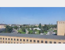 Blick von oben auf eine städtische Umgebung mit Gebäuden, einer Straße und einer Grünanlage mit Bäumen. Am Horizont sind der Berliner Fernsehturm und andere markante Gebäude sichtbar, bei klarem Himmel.