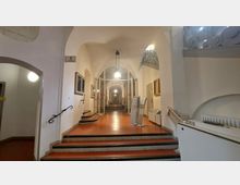 Ein Innenraum mit einem Korridor und Treppenstufen aus rotem Bodenbelag, umgeben von weißen Wänden und Rundbögen. Der Gang ist durch Hängelampen beleuchtet, an den Seiten befinden sich Türen, Geländer und Informationstafeln.