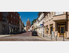 Straßenszene mit gepflasterter Fahrbahn, flankiert von mehrstöckigen historischen Gebäuden mit beige- und erdfarbenen Fassaden. Im Hintergrund ist ein mittelalterliches Backsteintor mit spitz zulaufendem Turm sichtbar; geparkte Autos und vereinzelte Laternen säumen die Straße.