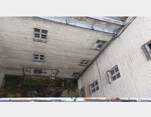 Blick in einen engen Innenhof umgeben von hohen, weiß-gemauerten Backsteinwänden mit kleinen, quadratischen Fenstern. Am Boden sind Gras, Moos und verstreute Blätter sichtbar, teilweise mit rostendem Metallgerüst im unteren Bereich.